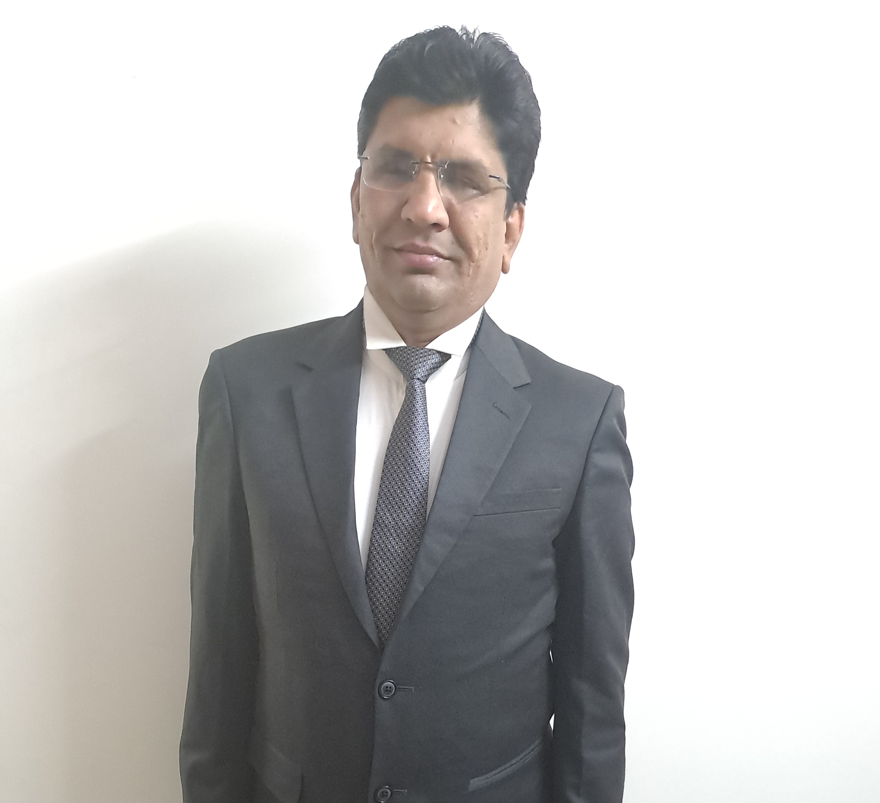 Akhilesh Malani wearing a dark charcoal suit, white dress shirt and grey patterned tie, standing against a white background. He has dark hair and wears thin-framed glasses.